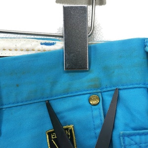 May include: Close-up of a pair of blue denim trousers with a gold button and a black label with the text "E.N.Y." on the front. The trousers have a light blue colour and a worn look.