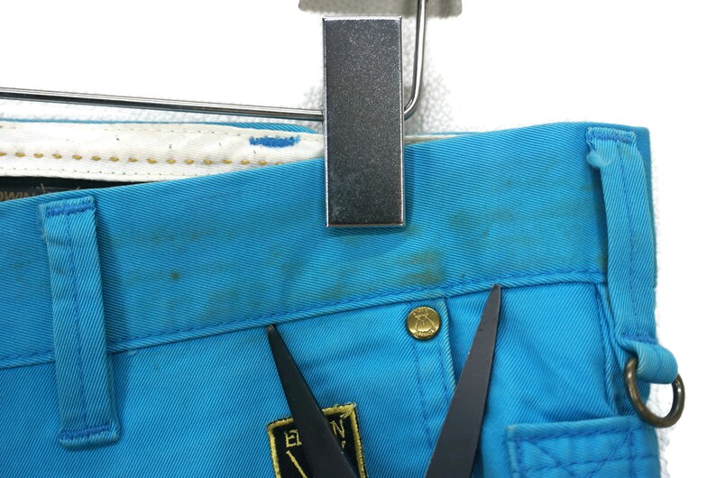 May include: Close-up of a pair of blue denim trousers with a gold button and a black label with the text "E.N.Y." on the front. The trousers have a light blue colour and a worn look.