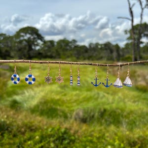 May include: A collection of nautical-themed earrings hanging from a twig. The earrings feature designs such as life preservers, ship wheels, anchors, and sailboats. The earrings are gold-toned with blue and white enamel details, set against a blurred green background.