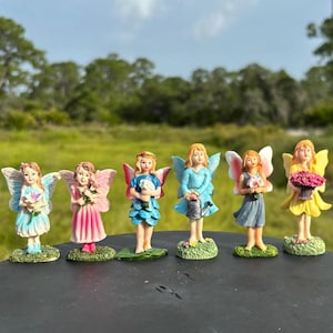 May include: Six miniature fairy figurines with colorful wings and dresses are arranged on a dark surface. Each fairy holds a different item, such as flowers or a watering can. The background features a green field and trees.