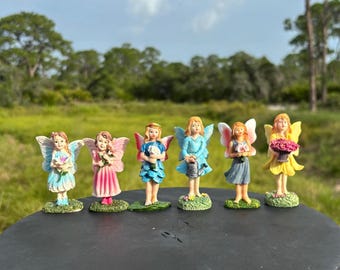 May include: Six miniature fairy figurines with colorful wings and dresses are arranged on a dark surface. Each fairy holds a different item, such as flowers or a watering can. The background features a green field and trees.