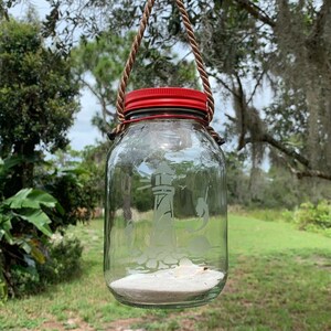 May include: A clear glass jar with a red lid and a rope handle, hanging from a tree branch. The jar features an etched lighthouse design, sand, and seashells inside. The background shows green foliage and grass.