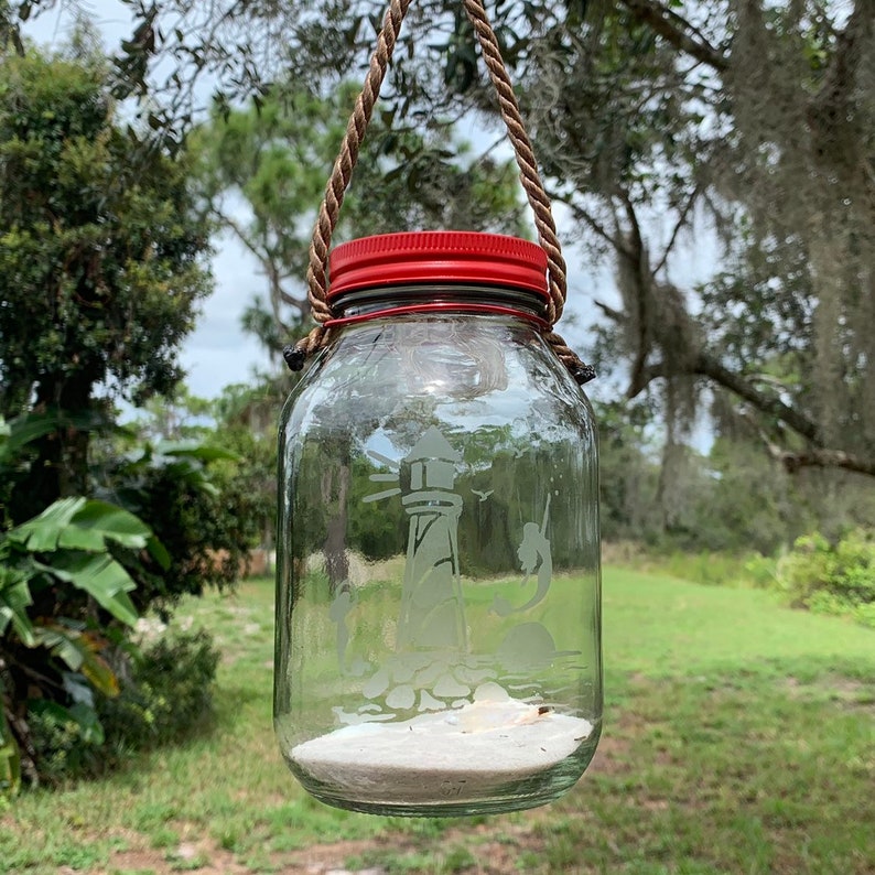 May include: A clear glass jar with a red lid and a rope handle, hanging from a tree branch. The jar features an etched lighthouse design, sand, and seashells inside. The background shows green foliage and grass.