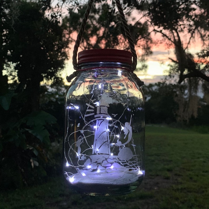 May include: A clear glass jar lantern with a red lid, etched with a lighthouse and figures, and filled with white fairy lights. The jar hangs from a rope handle against a dusk sky.