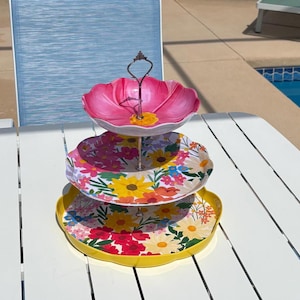 May include: A three-tiered serving tray with a floral design. The top tier is shaped like a pink flower, while the other two tiers are round with colorful flower patterns. The tray has a metal handle and is placed on a white table.