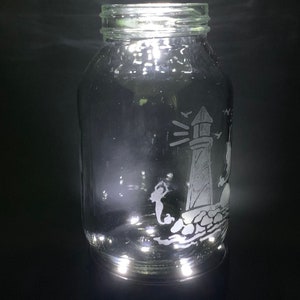 May include: A clear glass jar with a silver lid. The jar features an etched design of a lighthouse, a mermaid, and birds. The design is in white against the clear glass. The jar is cylindrical and sits on a dark surface.