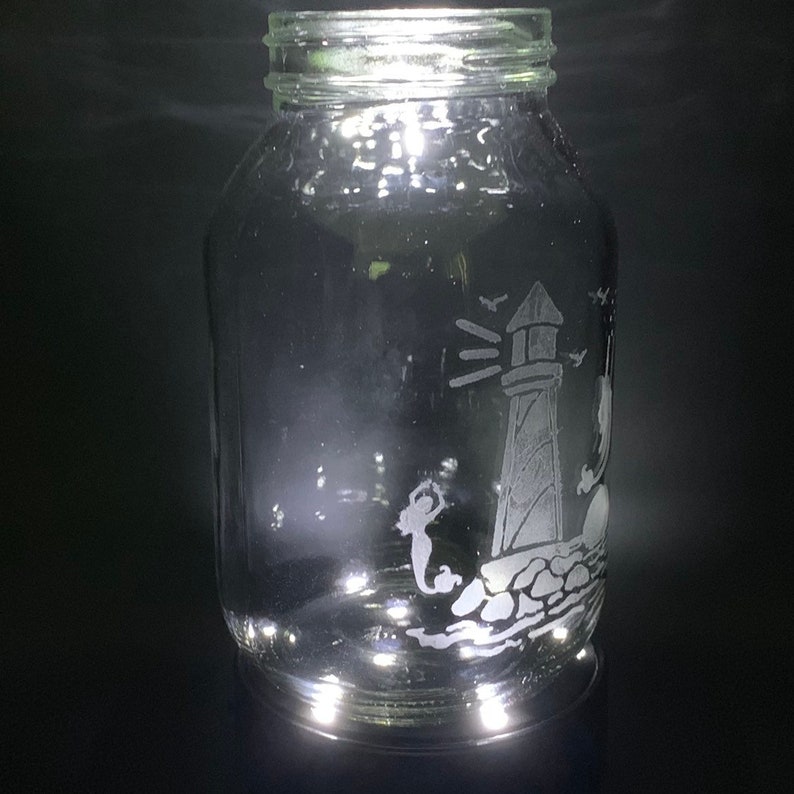 May include: A clear glass jar with a silver lid. The jar features an etched design of a lighthouse, a mermaid, and birds. The design is in white against the clear glass. The jar is cylindrical and sits on a dark surface.