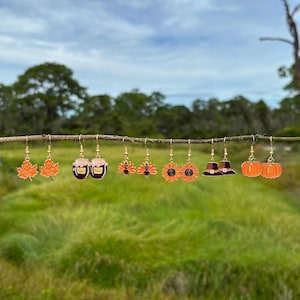 May include: A collection of autumn-themed earrings, including maple leaves, jam jars, turkeys, sunflowers, pilgrim hats, and pumpkins. Each earring is gold-toned with enamel detailing in shades of orange, brown, and black. The earrings are displayed on a thin branch against a blurred green background.