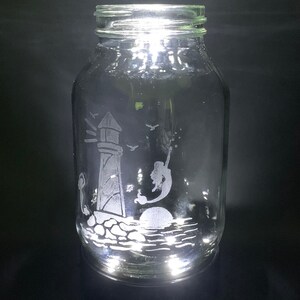 May include: A clear glass jar with a lighthouse, mermaid, and ocean scene etched on the front. The jar is illuminated from within, creating a glowing effect. The design includes a lighthouse, a mermaid, birds, and the sun.