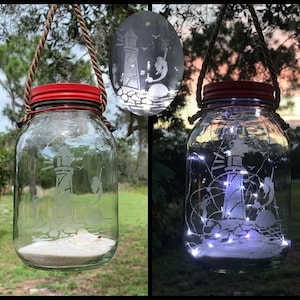 May include: Clear glass mason jar lantern with a red lid, featuring an etched lighthouse design. The jar hangs from a rope handle and is filled with sand. A smaller, similar lantern is also visible. The lantern is lit with white fairy lights.
