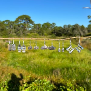 Puede incluir: Una colección de pendientes plateados colgados de una rama de madera. Los pendientes presentan varios diseños, incluyendo cámaras, barcos y formas abstractas. El fondo es una vista borrosa de hierba verde y árboles bajo un cielo azul brillante.