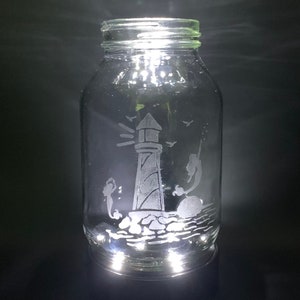 May include: A clear glass jar with a lighthouse and mermaid design etched on the surface. The lighthouse is on a rocky base, with mermaids and birds. The jar is illuminated from within, creating a glowing effect.