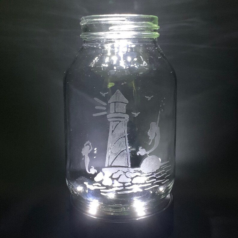 May include: A clear glass jar with a lighthouse and mermaid design etched on the surface. The lighthouse is on a rocky base, with mermaids and birds. The jar is illuminated from within, creating a glowing effect.