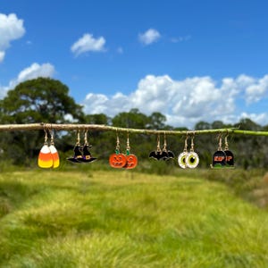 May include: A collection of Halloween-themed dangle earrings. The earrings include designs of a candy corn, witch hat, pumpkins, bat, eyeball, and a tombstone. Each earring is gold-toned with colorful enamel details, displayed on a natural branch against a blurred green and blue background.