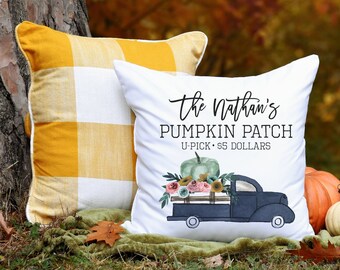 May include: White pillow with a black truck carrying pumpkins and flowers. The text on the pillow reads "The Nathan's Pumpkin Patch U-Pick $5 Dollars". The pillow is sitting on a green blanket with two pumpkins in the foreground.