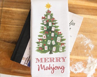 May include: White kitchen towel with a Christmas tree design featuring mahjong tiles, red bows, and a gold star. The words "MERRY Mahjong" are printed in red. The towel is on a wooden cutting board.