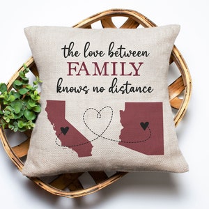 May include: A beige throw pillow with the text "the love between FAMILY knows no distance" printed in black. The pillow features an outline of California and Arizona in maroon with a heart connecting the two states.