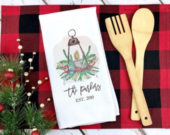 May include: White kitchen towel with a watercolor illustration of a lantern with a lit candle inside, surrounded by pine branches and berries. The text "The Parkers EST. 2010" is printed below the illustration. The towel is on a red and black buffalo plaid placemat.