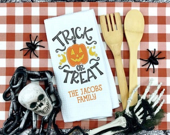May include: White kitchen towel with a black and orange pumpkin design and the text "Trick or Treat The Jacobs Family". The towel is on a red and white checkered tablecloth with a skeleton hand, a chain, and two spiders.