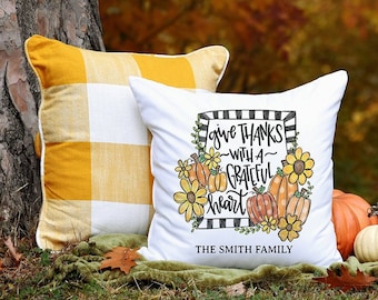 May include: White throw pillow with a black and white striped frame and the text "give thanks -with a grateful heart" with a watercolor illustration of sunflowers and pumpkins. The pillow also has the text "The Smith Family" at the bottom.