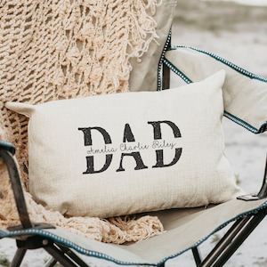 May include: A beige pillow with the word "DAD" printed in black with a distressed look. The names "Amelia, Charlie, Riley" are printed in black script above a black line under the word "DAD".