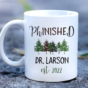 May include: White ceramic coffee mug with the text "Ph.inished" in black cursive font, a row of five green, brown, and black trees, the text "Dr. Larson" in black, and the text "est. 2022" in green.