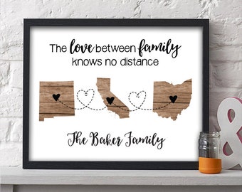 May include: A framed print with a woodgrain background featuring the text "The love between family knows no distance" and three states outlined in woodgrain with hearts connecting them. The states are New Mexico, California, and Ohio. The text "The Baker Family" is printed below the states.