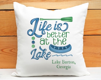 May include: White square pillow with the text "Life is better at the Lake" in blue and green script. The design includes a blue canoe, green oar, and the location "Lake Burton, Georgia".