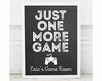 Pode incluir: Um sinal preto e branco com o texto "Just One More Game" e uma silhueta branca de um controlador de videogame. O sinal também diz "Eric's Game Room" na parte inferior.