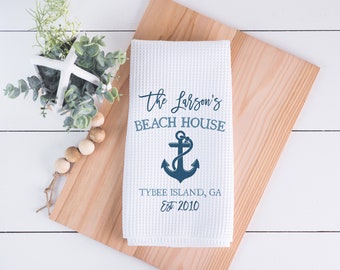 Könnte beinhalten: Weißes Küchenhandtuch mit Waffelstruktur und blauem Text "The Larson's BEACH HOUSE" sowie einer Ankergrafik. Unter dem Anker steht "TYBEE ISLAND, GA EST 2010". Das Handtuch liegt auf einem Holzschneidebrett.