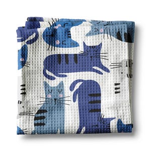 May include: A white waffle weave kitchen towel with a blue and white cat pattern. The cats are in various poses, including sleeping, sitting, and standing. The towel is perfect for drying dishes or wiping up spills.