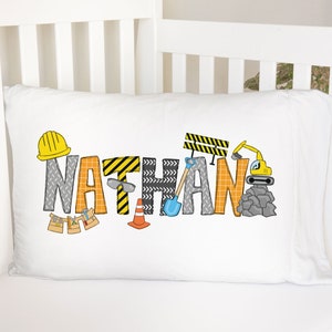 May include: A white pillowcase with the name "NATHAN" spelled out in colorful letters. The letters are decorated with construction-themed images, including a hard hat, a shovel, a traffic cone, and a construction vehicle.