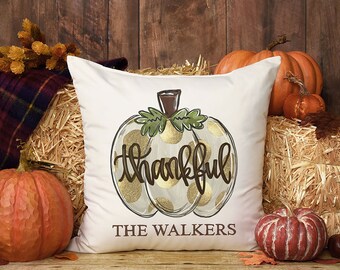 May include: White throw pillow with a gold and green pumpkin design. The pumpkin has gold polka dots and the word "thankful" written on it. The pillow also has the text "THE WALKERS" printed on it.