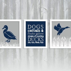 May include: Set of three framed prints with duck illustrations. One print features a duck standing, another a flying duck, and the third has text that reads "DOGS cattails & flocks galore dreams of counting DUCKS one, two, three, four". The ducks are navy blue on a gray background.