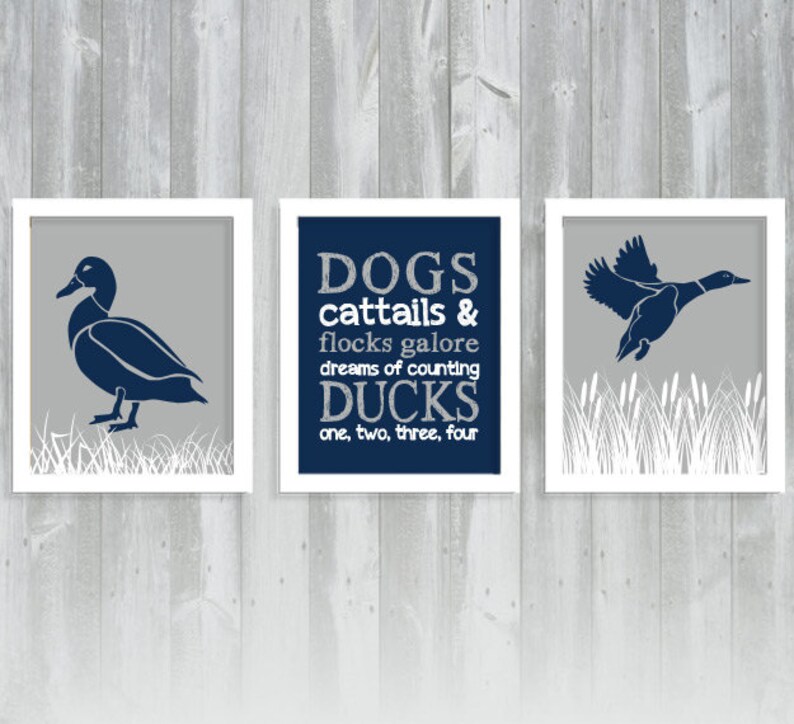 May include: Set of three framed prints with duck illustrations. One print features a duck standing, another a flying duck, and the third has text that reads "DOGS cattails & flocks galore dreams of counting DUCKS one, two, three, four". The ducks are navy blue on a gray background.