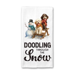 May include: White waffle-weave kitchen towel featuring a watercolor illustration of a snowman and two dogs on a wooden bench in the snow. The towel has the text "DOODLING THROUGH THE SNOW" in black script.