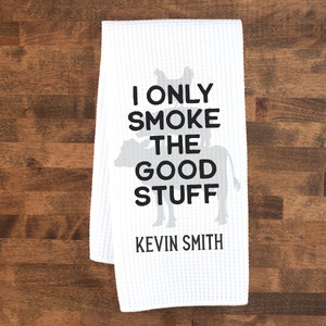 May include: White kitchen towel with a black silhouette of a cowboy riding a cow. The text reads "I only smoke the good stuff" and "Kevin Smith".
