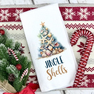 May include: White kitchen towel with a Christmas tree made of seashells and starfish. The text "Jingle Shells" is printed in blue and gold.