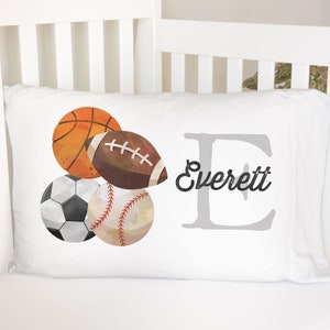May include: White pillowcase featuring watercolor illustrations of a basketball, football, soccer ball, and baseball. The name "Everett" is written in a script font next to a large letter "E". The pillow is placed on a white crib.