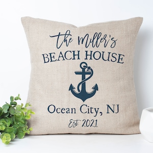 May include: A beige linen pillow with the text "The Miller's Beach House" in navy blue lettering.  Below the text is a navy blue anchor and the text "Ocean City, NJ Est. 2021".