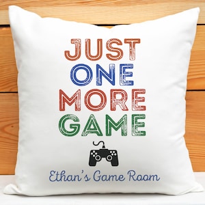 May include: White throw pillow with the text "JUST ONE MORE GAME" in colorful, distressed lettering. A black video game controller icon is below the text. The text "Ethan's Game Room" is at the bottom of the pillow.