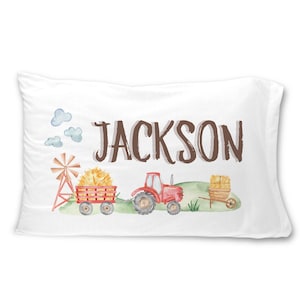 May include: White pillowcase with a watercolor farm scene. Features a red tractor, hay wagon, windmill, and the name "JACKSON" in brown letters. Includes blue clouds and green grass.