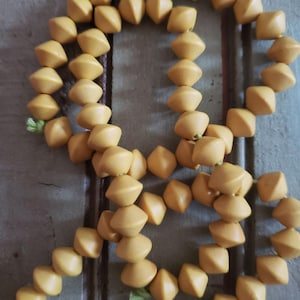 May include: A necklace composed of numerous yellow, geometric beads. The beads are diamond-shaped with rounded edges, strung together with a green string. The necklace is displayed against a weathered, light blue-green surface.