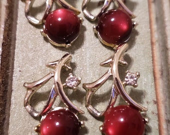 May include: Four gold-toned earrings with a leaf-like design. Each earring features a large, round, deep red stone and a small, clear gemstone accent. The earrings are displayed on a textured, light green surface.