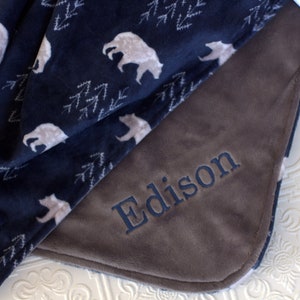 Op de afbeelding: Een gepersonaliseerde blauwe en bruine fleece deken met een witte berenprint en de naam "Edison" geborduurd op de bruine kant.