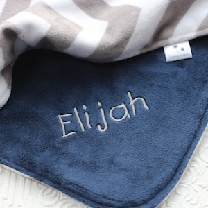 May include: A soft, blue baby blanket with a gray and white chevron pattern on the reverse side. The blanket is personalized with the name "Elijah" embroidered in white thread.