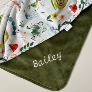 May include: A green and white baby blanket with a whimsical woodland design featuring frogs, dragonflies, and mushrooms. The blanket is personalized with the name "Bailey" embroidered in white on the green side.