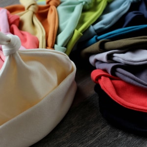 May include: A collection of colorful fabric hats with a knotted design. The hats are made of soft, stretchy material and come in a variety of colors, including pink, yellow, orange, green, blue, and gray.