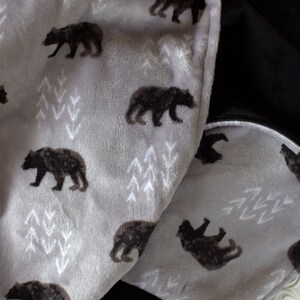 May include: A soft grey and black fleece blanket with a repeating pattern of black bears and white stylised trees. The blanket is folded in half, showing the grey side with the pattern and the black side.