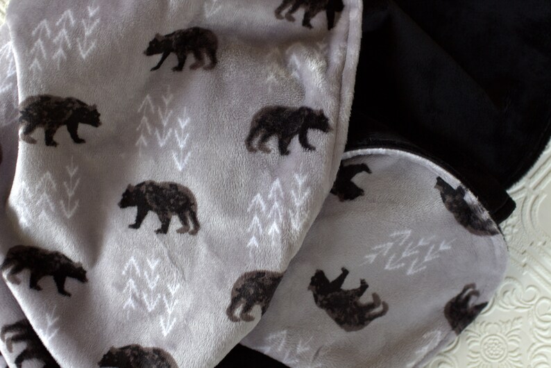 May include: A soft grey and black fleece blanket with a repeating pattern of black bears and white stylised trees. The blanket is folded in half, showing the grey side with the pattern and the black side.
